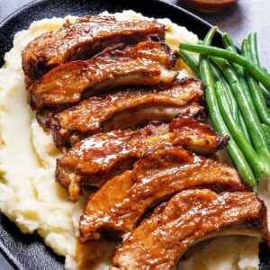 Slow-cooked barbecue ribs resting on a bed of mashed potatoes, accompanied by a side of green beans, served on a black serving plate.