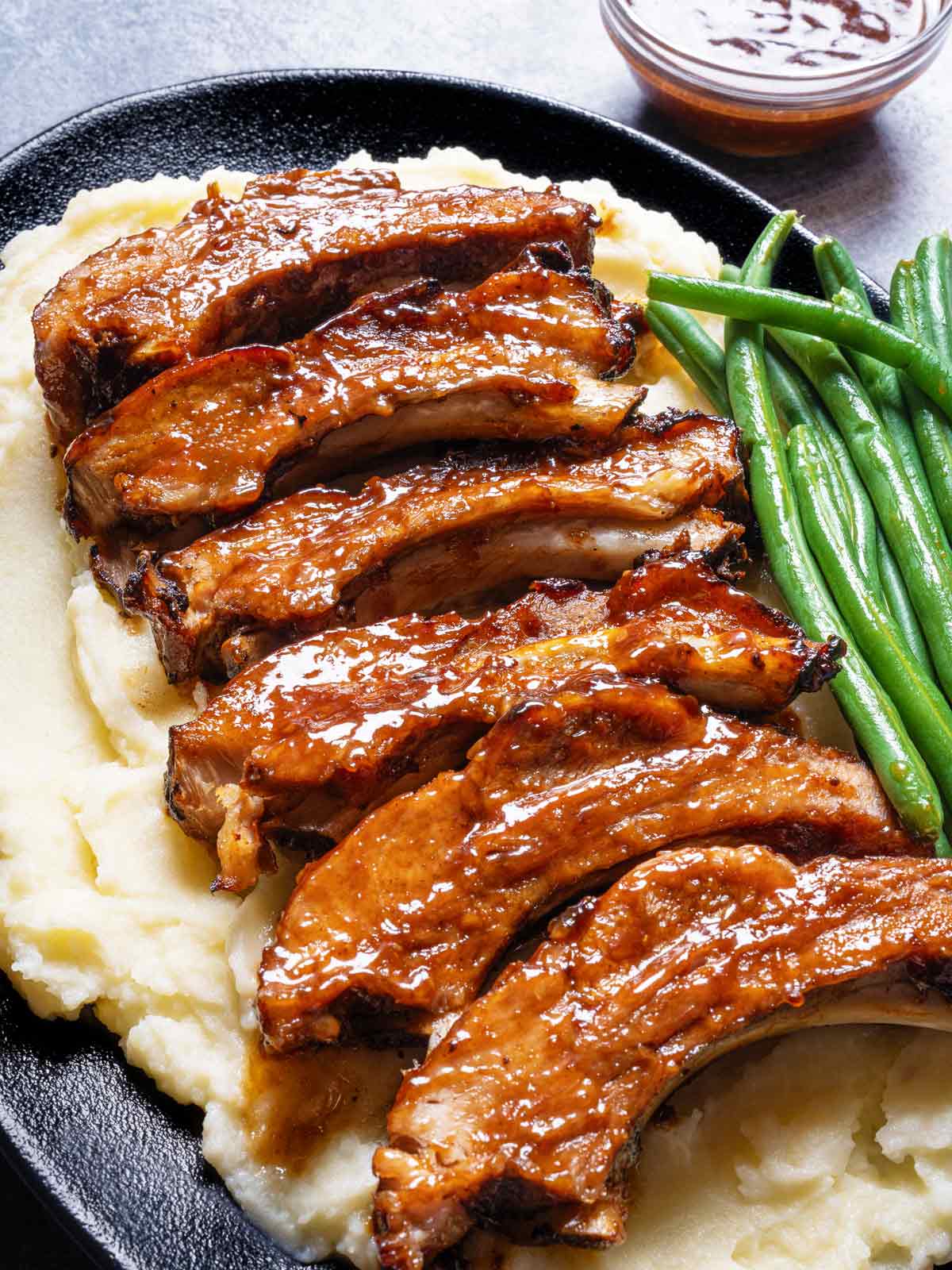 Slow-cooked barbecue ribs resting on a bed of mashed potatoes, accompanied by a side of green beans, served on a black serving plate.