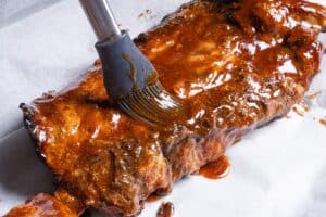 Rack of ribs being glazed with barbecue mixture on a baking tray.