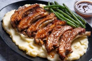 Barbecue slow cooker ribs cut individually, placed on mashed potato with beans on the side, and barbecue sauce next to the plate.