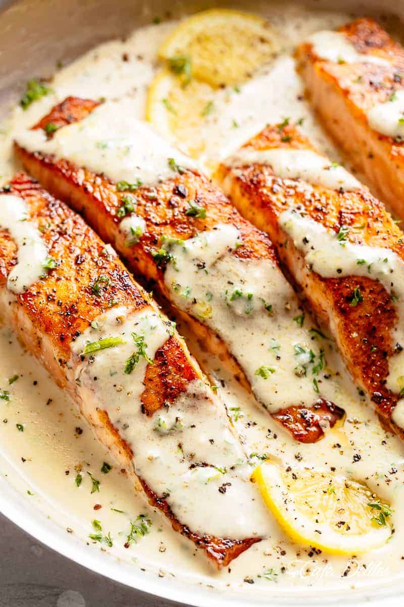 Four soft salmon fillets sitting and covered in creamy garlic butter sauce. Fresh herbs and two lemon wedges are scattered across the top of the fish.