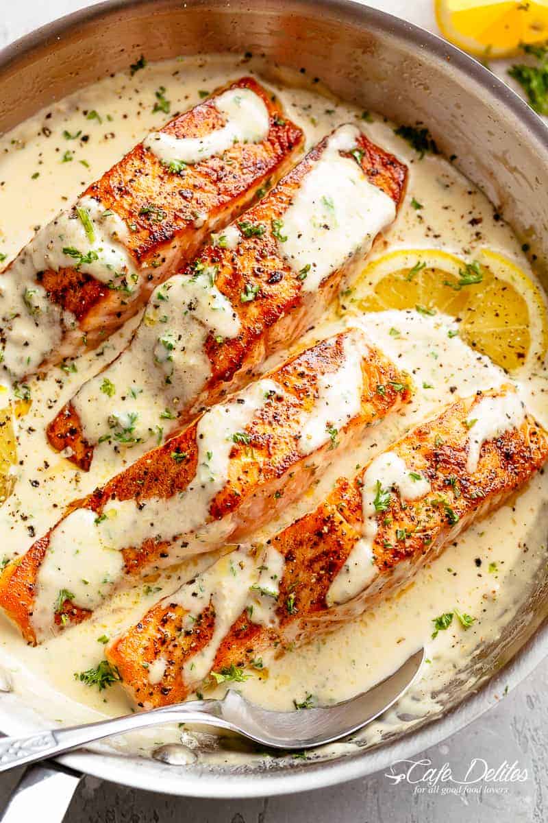 Four salmon fillets in a creamy garlic butter sauce in a large pan with a serving spoon resting inside the pan. Fresh green herbs are sprinkled over top of the fish and two lemon wedges sit beneath two of the fillets, looks yummy !