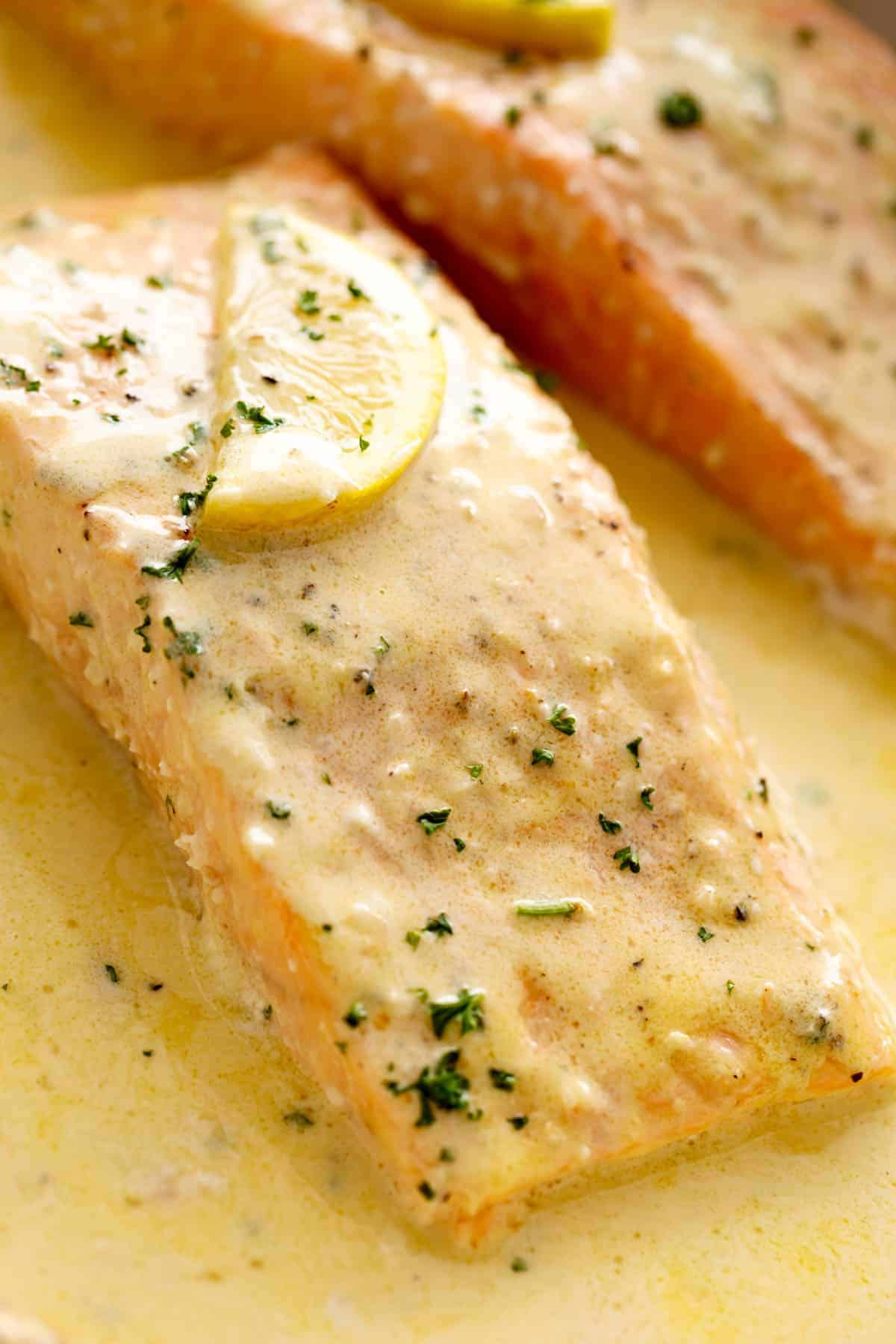 Close up image of salmon piece in lemon butter sauce with slice of lemon and chopped herbs on top.