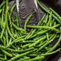 Sautéed, buttery green beans covered in garlic with tongs leaning on side of pan.