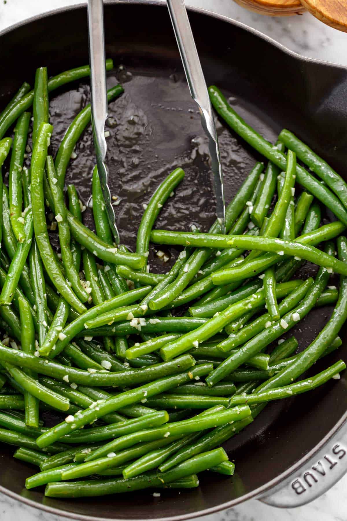 Sautéed, buttery green beans covered in garlic with tongs leaning on side of pan.