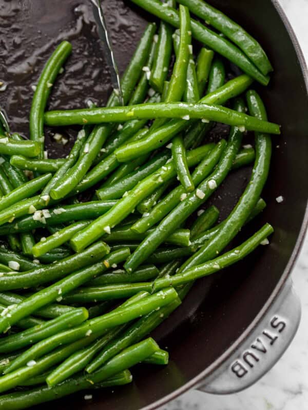 Close up image of Sautéed green beans with a sprinkle of course salt in a pan being pushed aside by a pair of tongs.