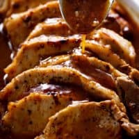 Side view image of A thick brown gravy being poured over thick slivers of pork loin roast with a large silver tablespoon.
