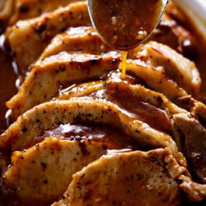 Side view image of A thick brown gravy being poured over thick slivers of pork loin roast with a large silver tablespoon.