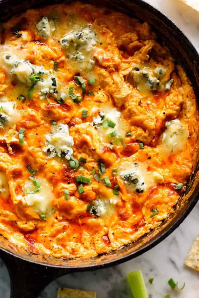 This close-up shot shows a cast iron skillet filled with a creamy, cheesy buffalo chicken dip. The dip is a vibrant orange color, with melted cheese, chunks of chicken, and green onion scallions sprinkled throughout. There are also generous dollops of blue cheese sprinkled on top. The edges of the skillet are darkened, suggesting the dip has been baked. The background is a marble surface, a few pieces of celery, and the corner of a cracker. Everything looks yummy