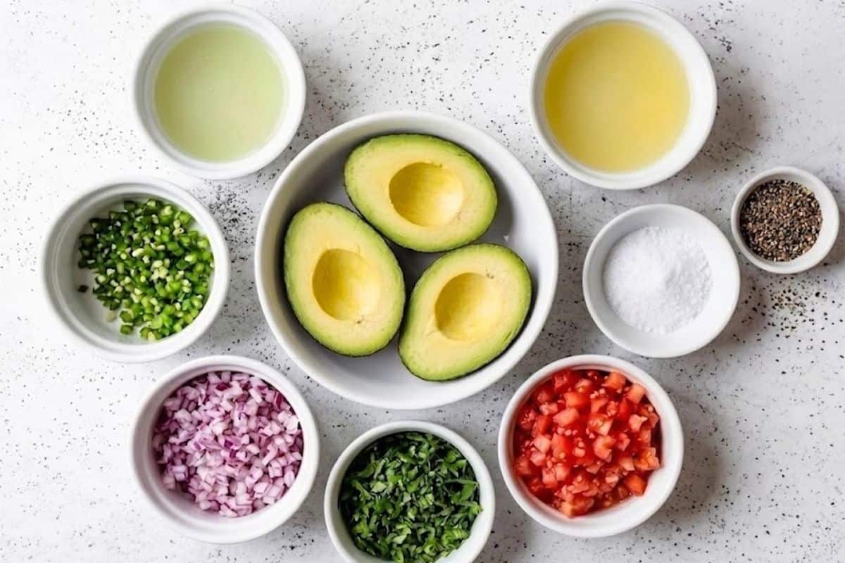 A flatlay image of labeled guacamole ingredients in small bowls arranged on a light surface. Visible ingredients include chopped red onion, diced tomato, green chile, chopped cilantro, salt, cracked black pepper, lime juice, lemon juice, and three halved ripe avocados in the center.