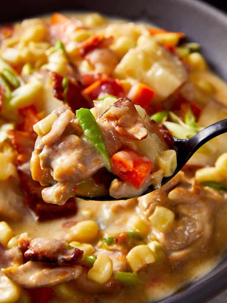 A white bowl of thick and creamy chicken corn chowder. A hand stirs the chowder with a tablespoon.