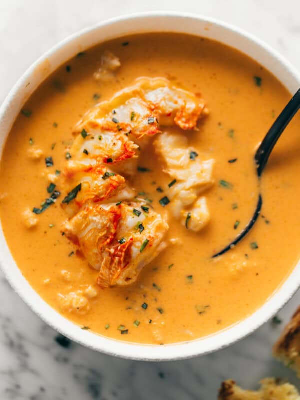 Cooked lobster flesh submerged in an orange soup in a white bowl. A black spoon is scooping some of the orange liquid in a bowl and spoon.