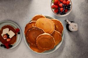served pancakes in a plate with maple syrup in a side, berries and a plate of served pancakes with butter, berries and maple syrup