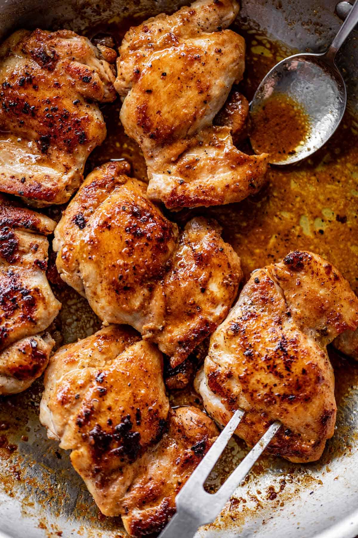 Crispy boneless chicken thighs in a silver pan, being bathed in their own jus with a spoon.