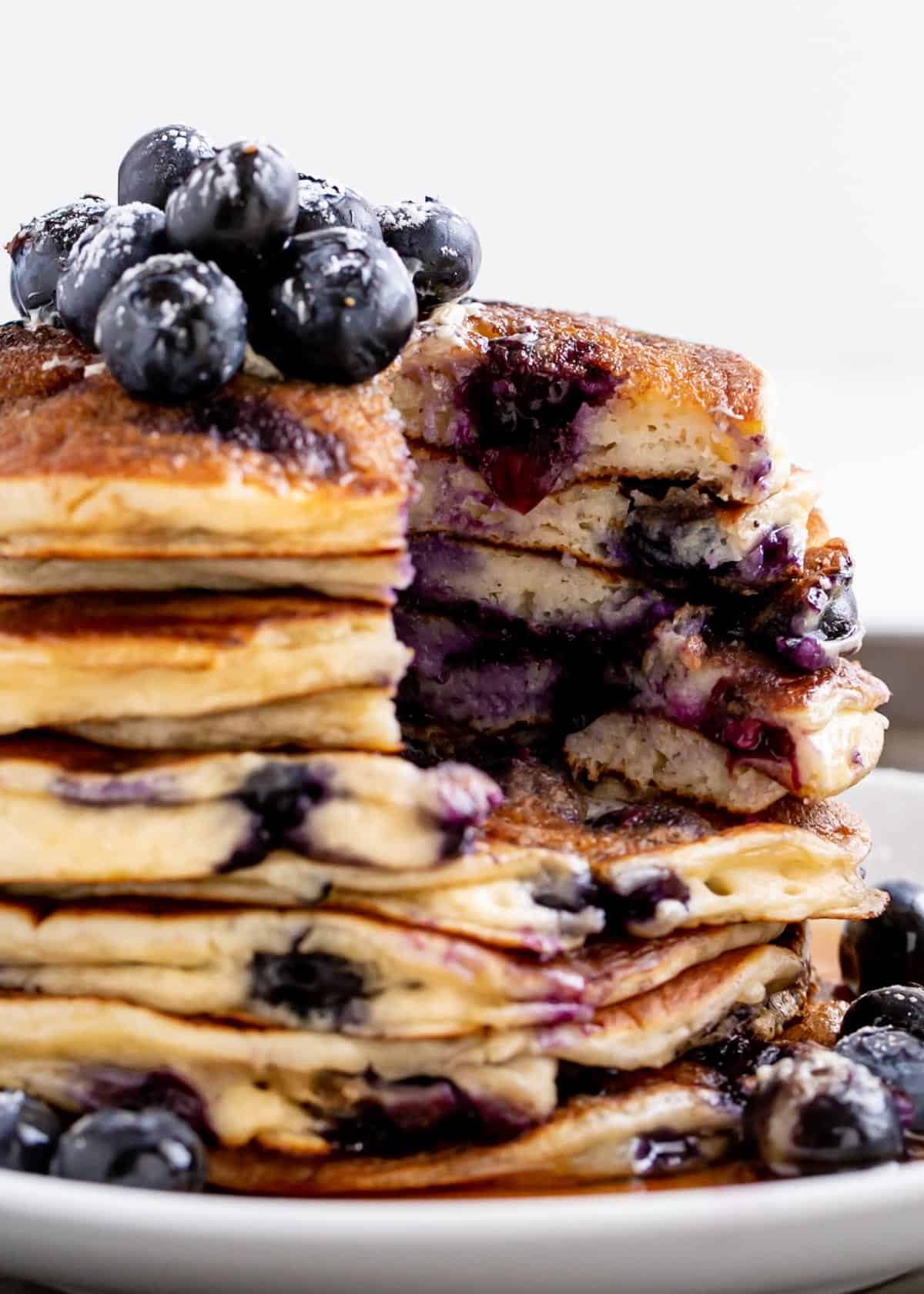 Cutting into a stack of Blueberry Ricotta Pancakes on a plate with maple syrup. Topped with fresh blueberries