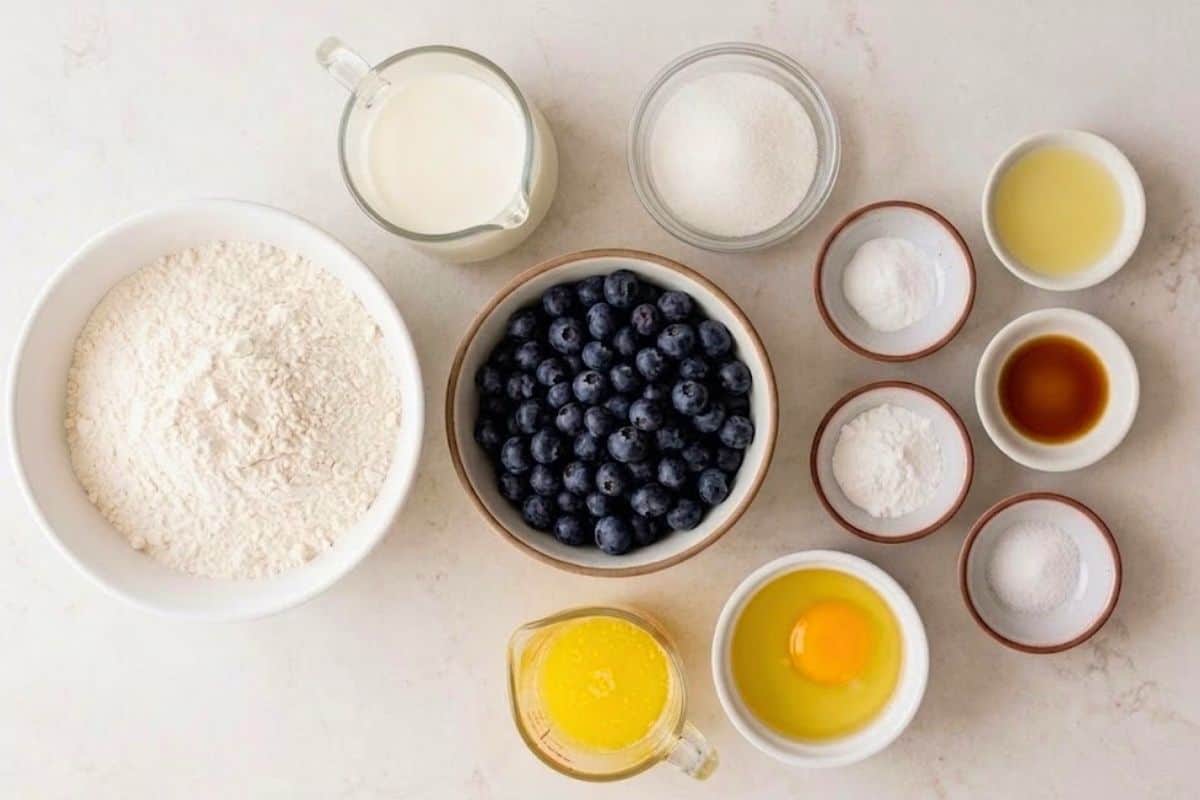 Overhead photo of labeled ingredients for Blueberry Pancakes, arranged in small bowls on a neutral surface. Labels include: Flour, Sugar, Baking Powder, Baking Soda, Salt, Milk, Butter, Vanilla, Lemon Juice, Egg, and Blueberries.