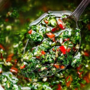 A small serving spoon lifts a spoonful of vibrant chimichurri, showcasing green herbs and red chili pieces mixed with oil from a bowl.
