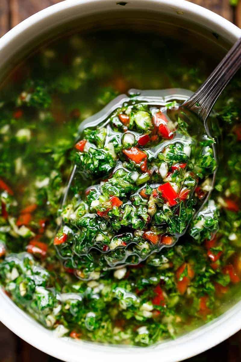 A small serving spoon lifts a spoonful of vibrant chimichurri, showcasing green herbs and red chili pieces mixed with oil from a bowl.