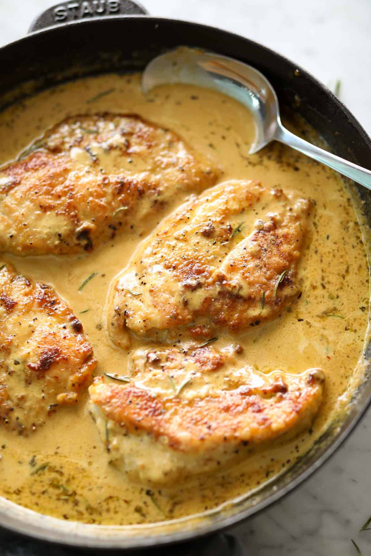 Creamy mustard chicken breasts pieces in a black Staub skillet with a metal spoon.