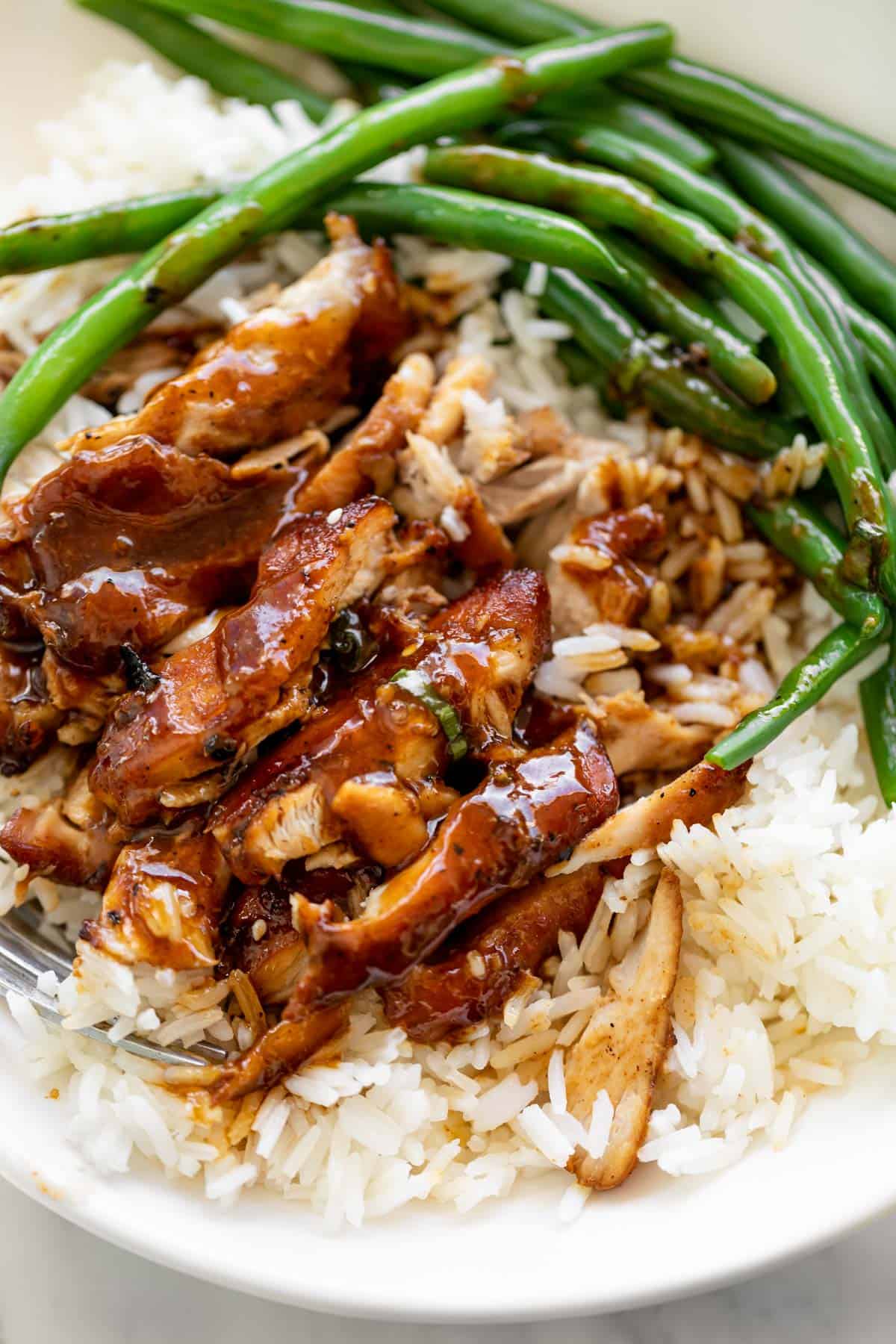Slow cooked honey garlic chicken sits on top of plain white rice in a white bowl with sautéed green beans.