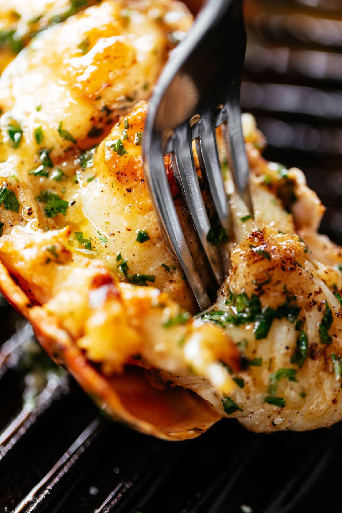 A fork pierces through a grilled lobster tail brushed with melted garlic herb butter on a black grill pan.