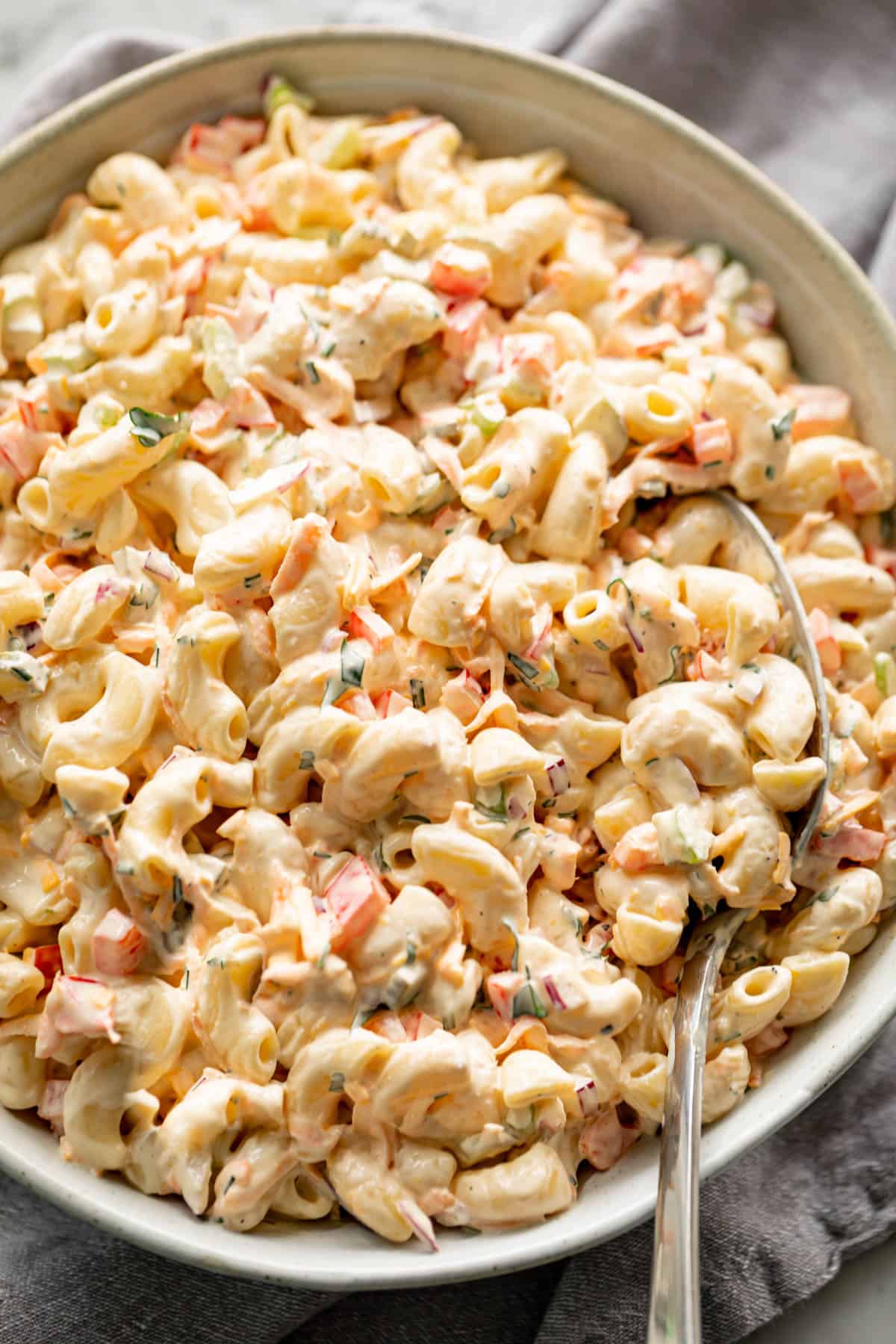 DELICIOUS! Macaroni Salad is quick to throw together and Served in a light grey bowl on a grey linen cloth with a spoon for scooping.