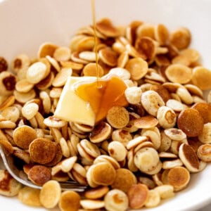 Mini Pancakes Cereal served with a piece of butter and drizzled with maple syrup. Served in a white bowl with a silver fork.