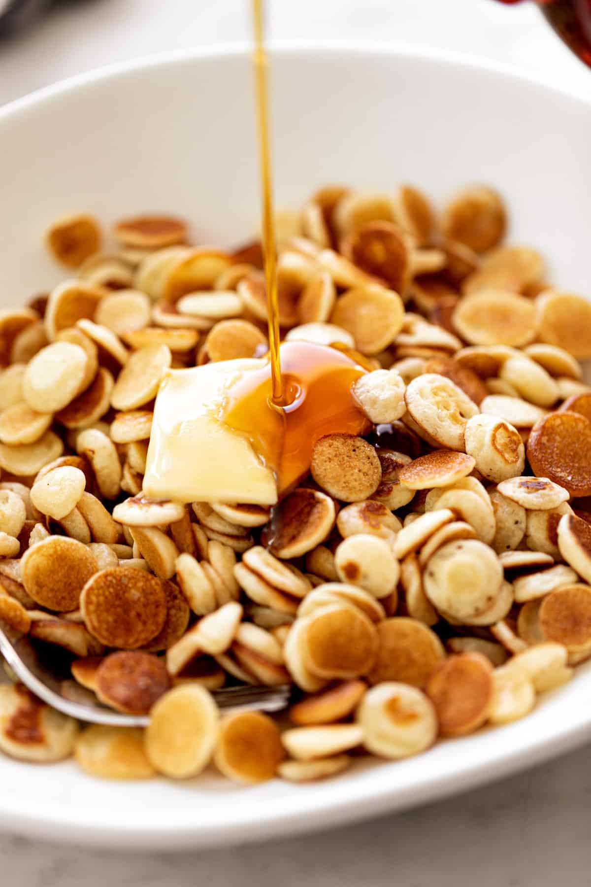 Maple syrup drizzled over Mini Pancakes Cereal served with a piece of butter in a white bowl with a silver fork.