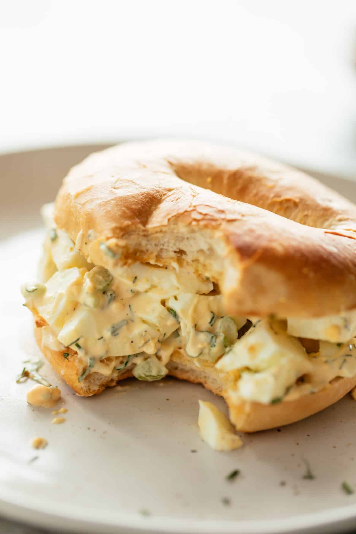 Perfect Egg Salad sandwiched in a bagel on a white plate sprinkled with fresh dill, chives and parsley.