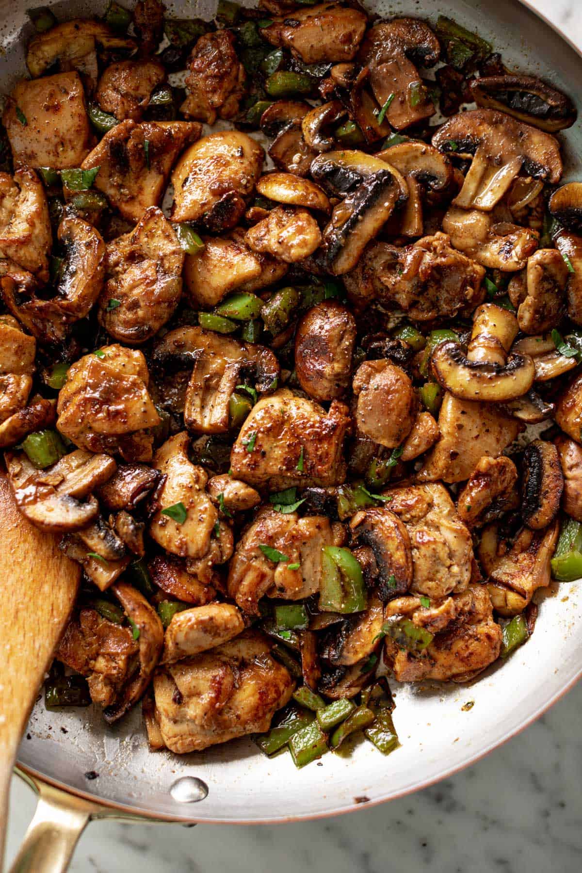 garlic mushroom chicken sautéing in a pan with a wooden spoon on the side.