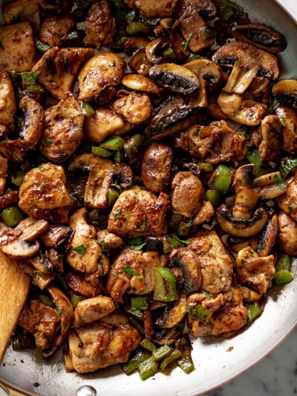 A wooden spoon stirring a pan filled with cooked mushrooms, chopped green onion and thick chicken pieces.