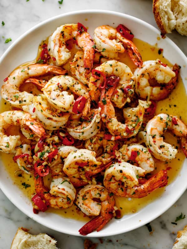 A pile of shrimp on a white plate sitting in chili garlic butter sauce. Served with broken bread pieces.