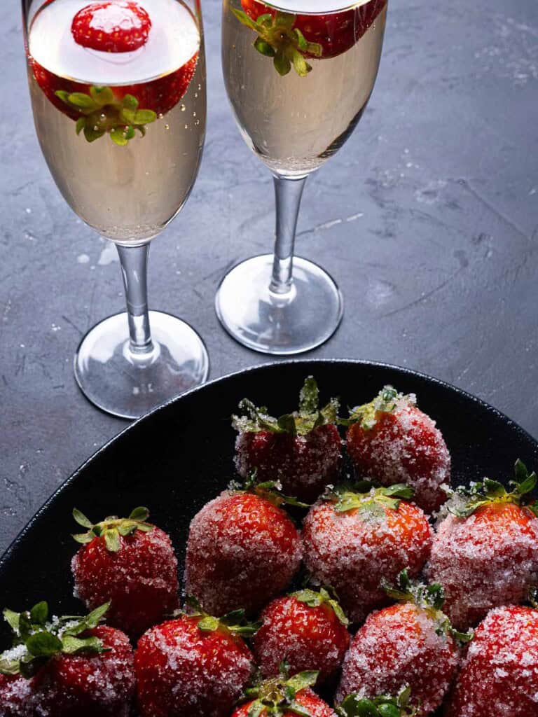 An image of strawberries coated in sugar and two flutes filled with champagne, each containing a strawberry.