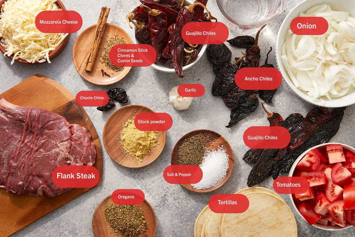 Flat lay of key birria taco ingredients. All labeled and arranged on a stone background.
