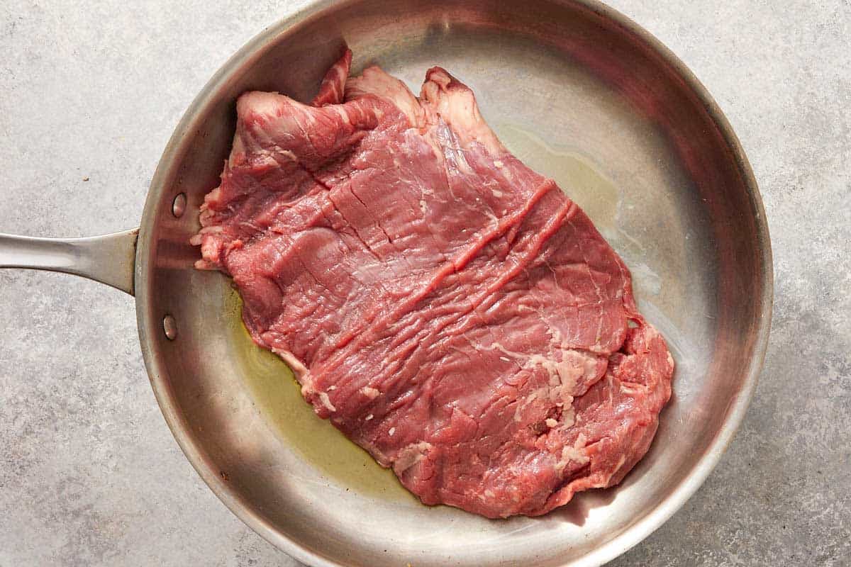 Raw flank steak searing in a stainless steel pan.