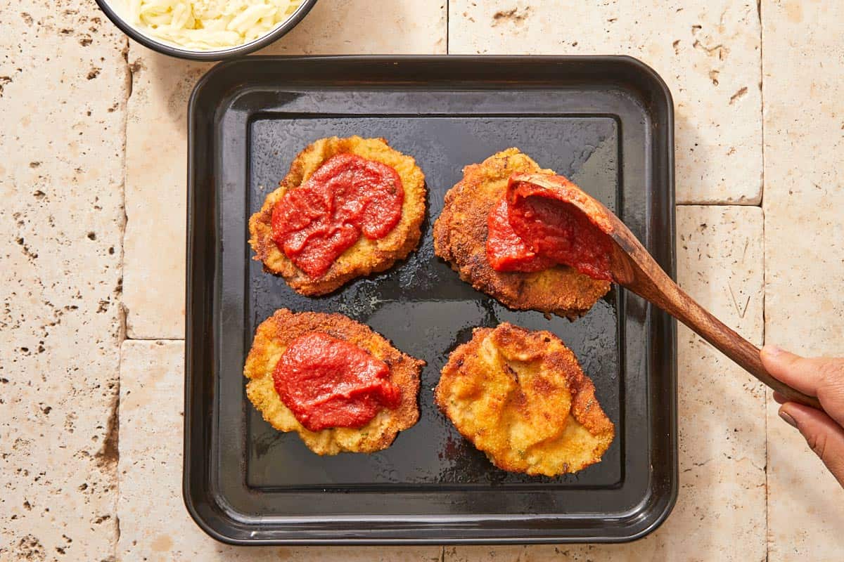 Hand spooning rich tomato sauce over crispy schnitzels