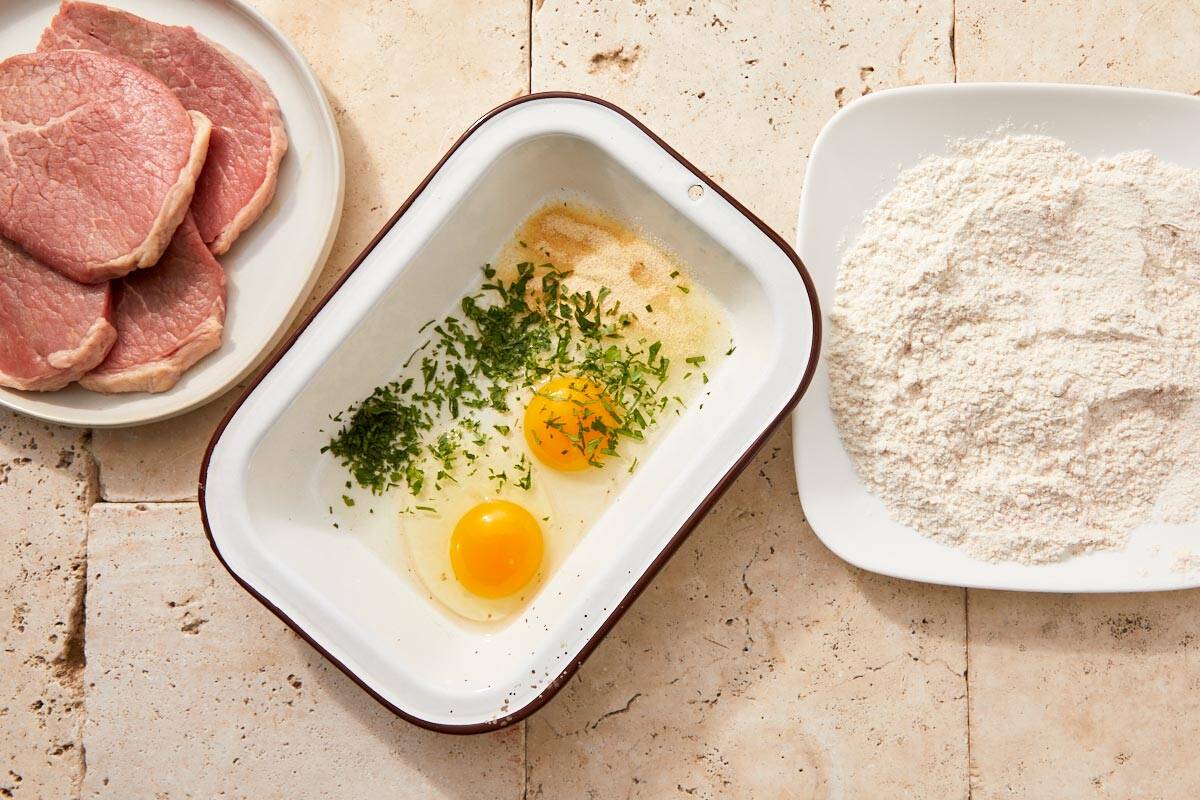 Raw beef slices next to a dish of flour with spices and a bowl of cracked eggs with chopped parsley and onion powder.