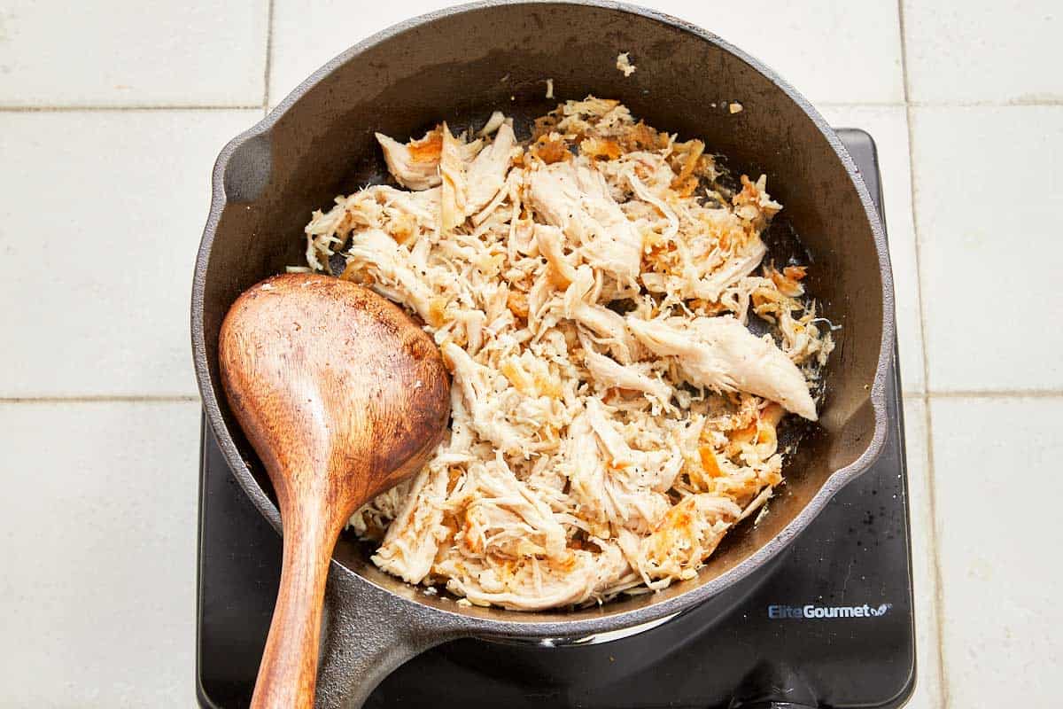 A skillet with sautéed shredded chicken and a wooden spoon.