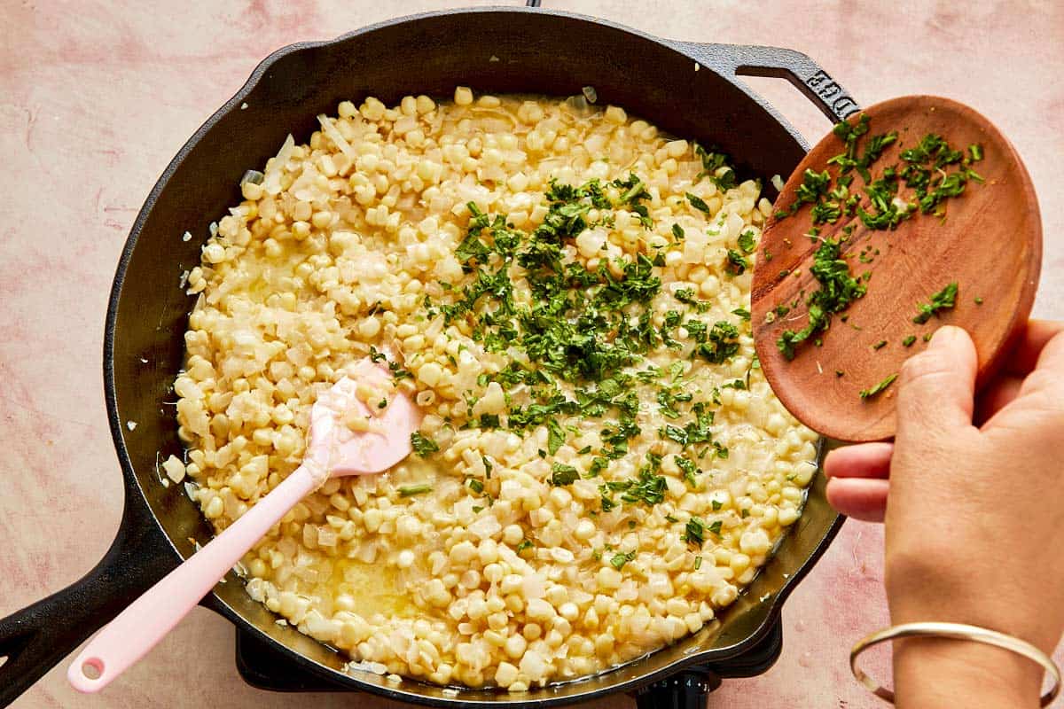 A person mixes corn in a skillet, ready to add chicken broth and epazote for simmering.