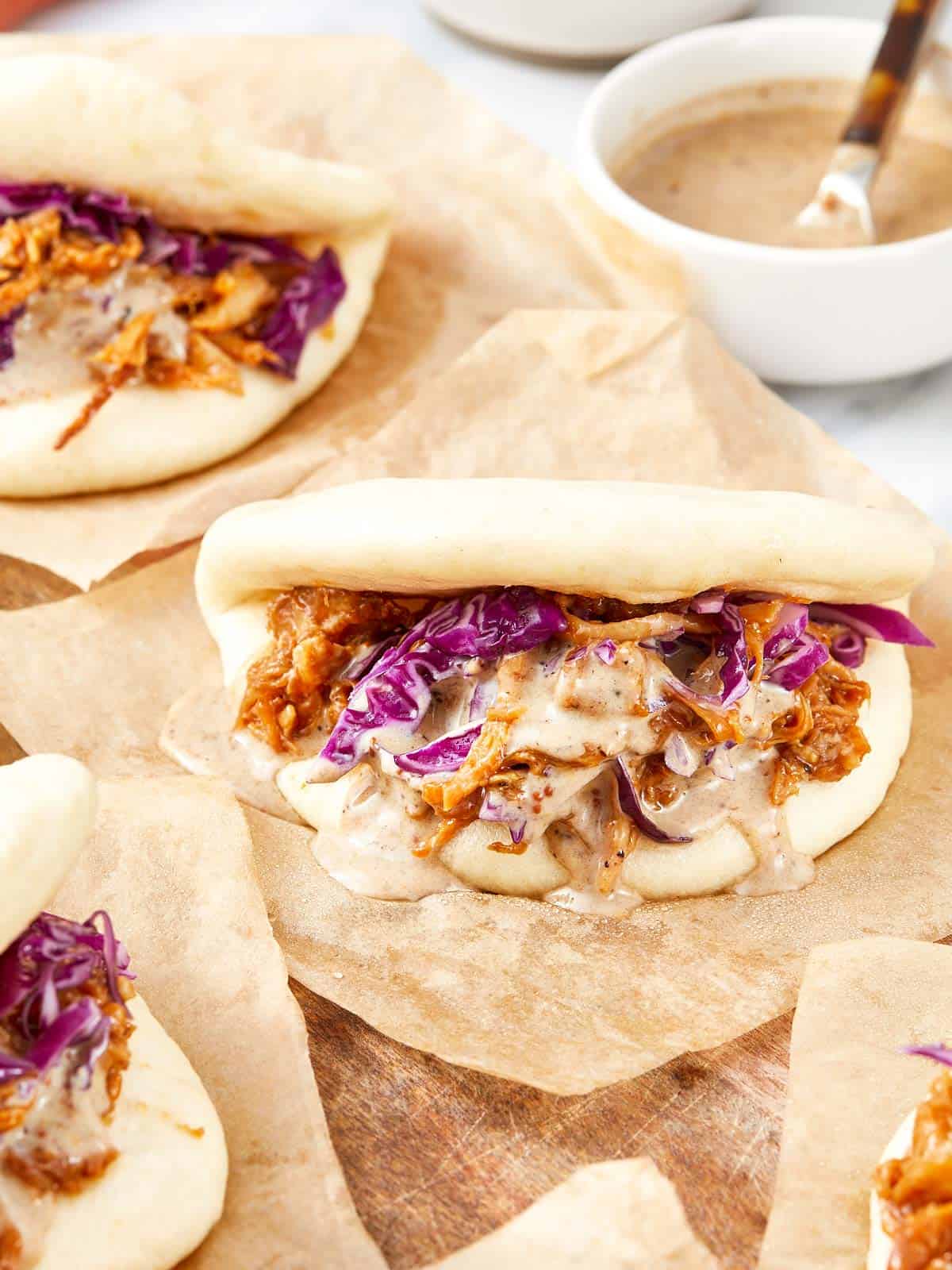 Close-Up of steamed bao buns filled with pulled pork, red cabbage, and drizzled black garlic mayo on parchment paper.