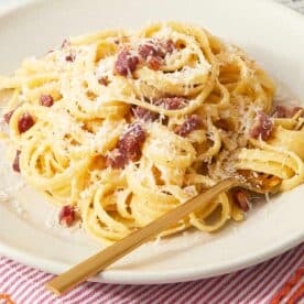 A plate of finished pasta carbonara on a red-striped napkin, topped with crispy pancetta and extra cheese.