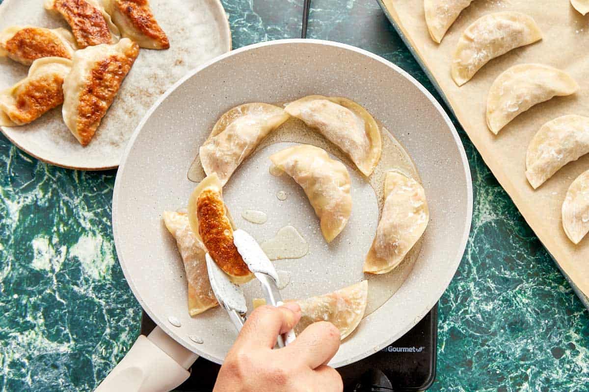 A person frying dumplings in a pan, steam rising as they add water and cover to cook until soft on top.