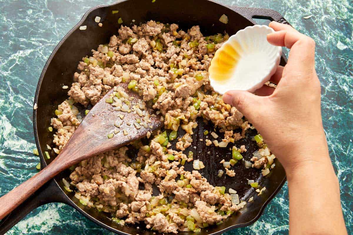 A person mixes ground meat in a skillet, deglazing with Shaoxing wine and soy sauce to enhance the dish's flavor.