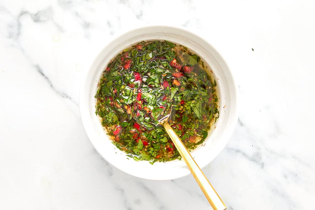 Chimichurri sauce fully mixed in a white bowl with visible herbs and chili, ready to rest.