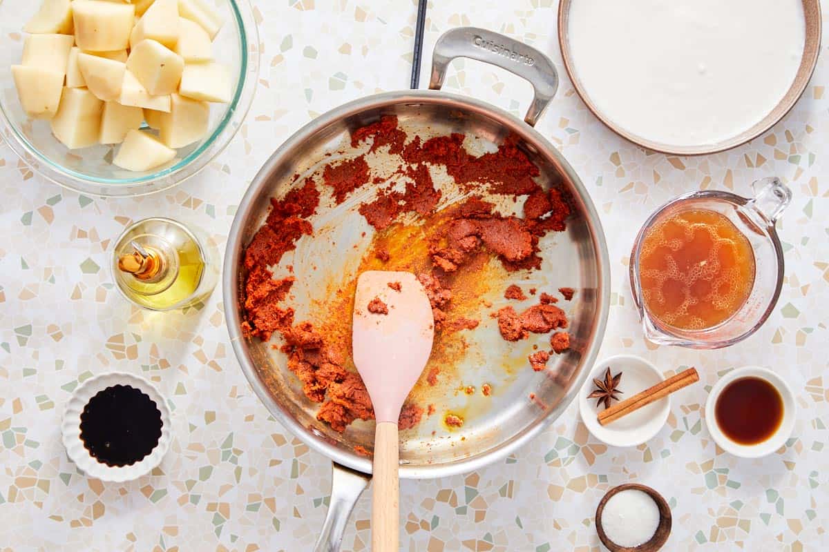 Curry paste cooking in a pan with potatoes, broth, and coconut milk nearby.