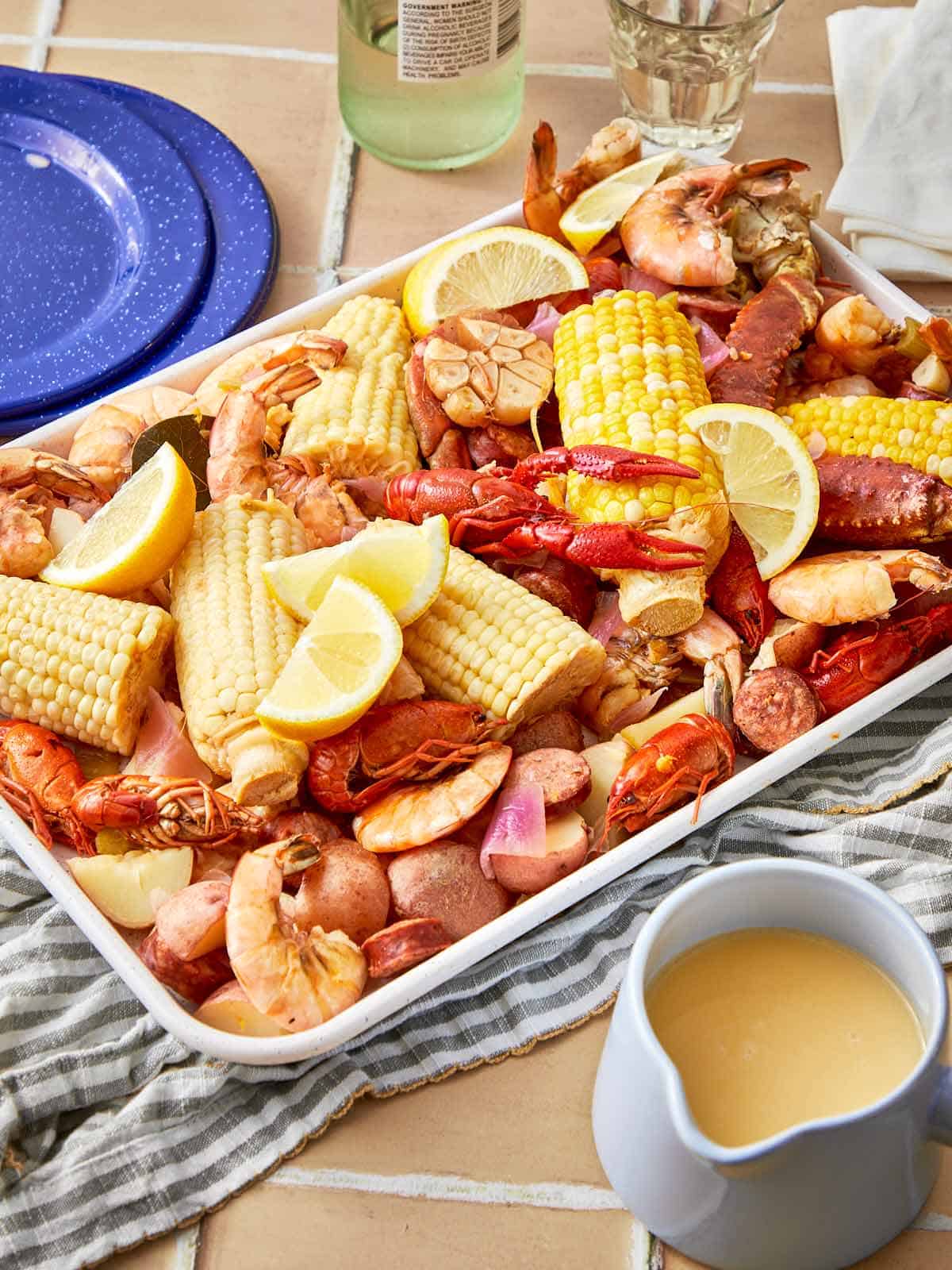 Seafood boil served on a white tray with corn, shrimp, crawfish, potatoes, lemon wedges, garlic, and sausage, with a side of buttery dipping sauce.
