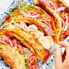 Close-up of smash burger tacos in a blue speckled tray, filled with melted cheese, lettuce, tomato, and red onion, being topped with creamy sauce.