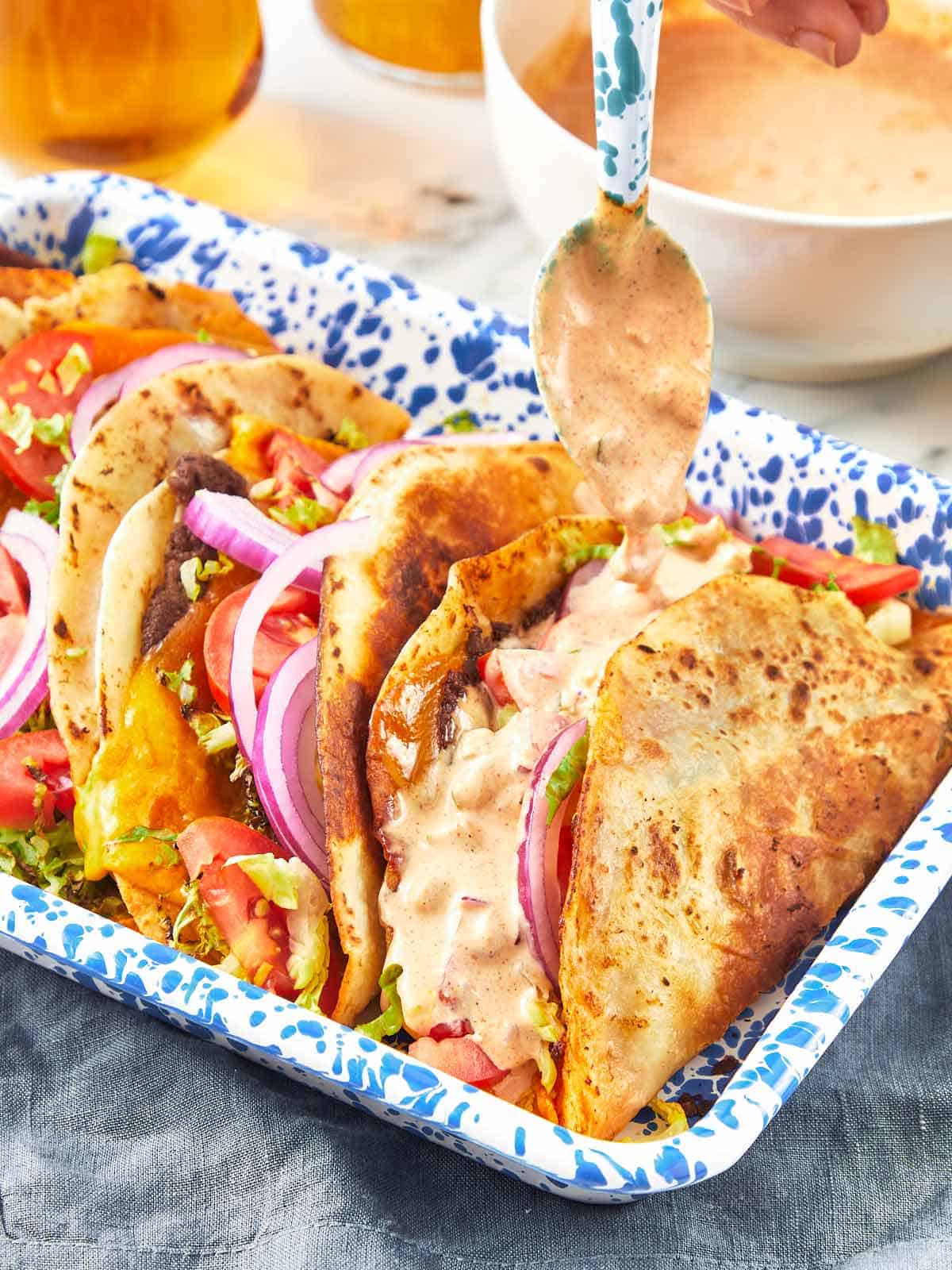 Close-up of smash burger tacos in a blue speckled tray, topped with lettuce, tomato, red onion, melted cheese, and drizzled with creamy sauce.