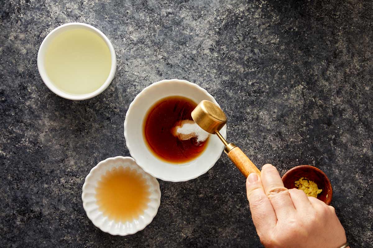 Hand adding sugar to soy sauce mixture in a white bowl with small bowls of vinegar and ginger.