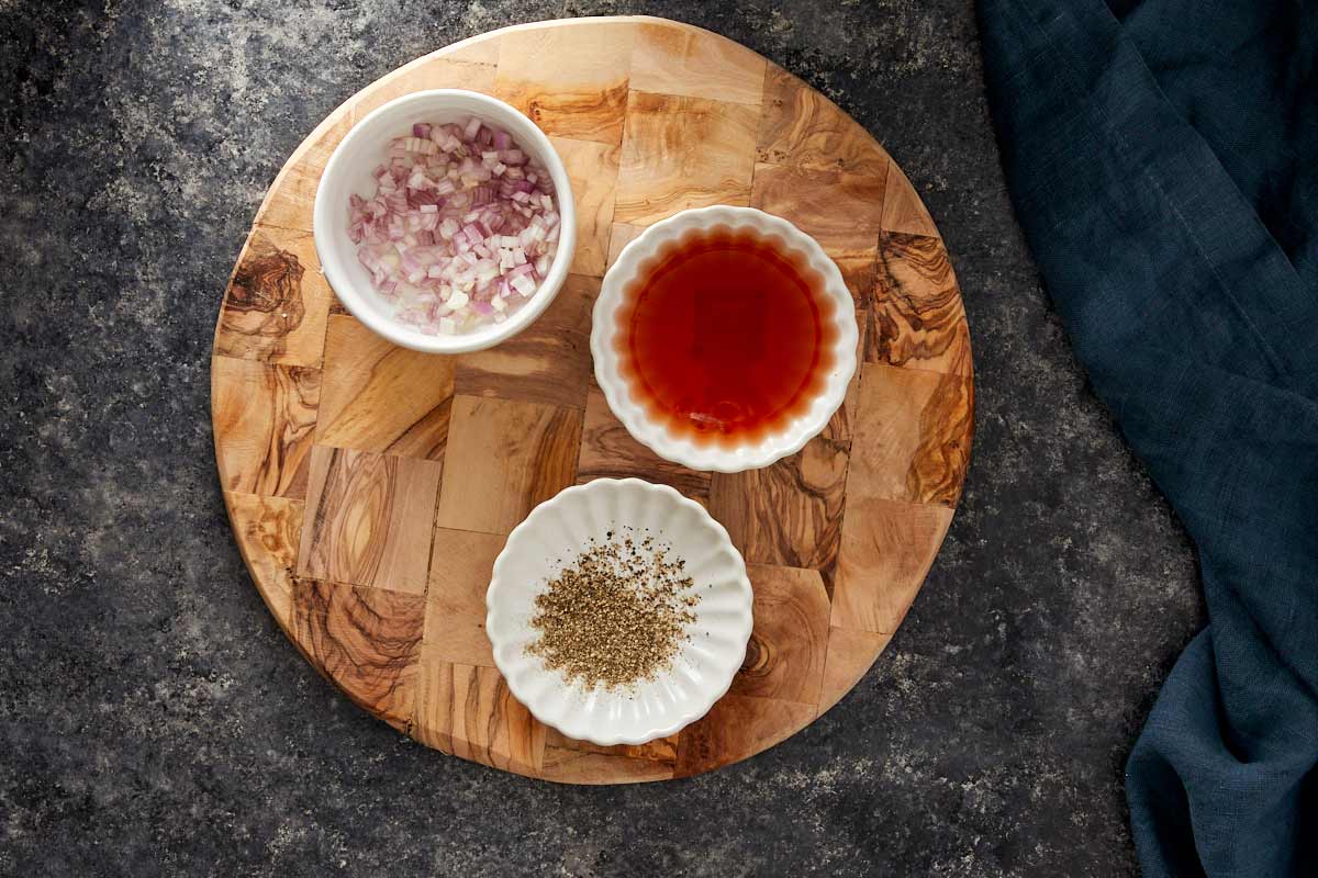 Small bowls with chopped shallots, red wine vinegar, and ground pepper on a wooden board.